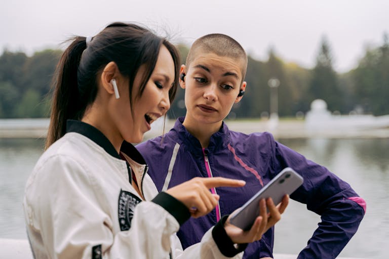 Two women in active wear enjoy music outdoors, sharing a smartphone experience.